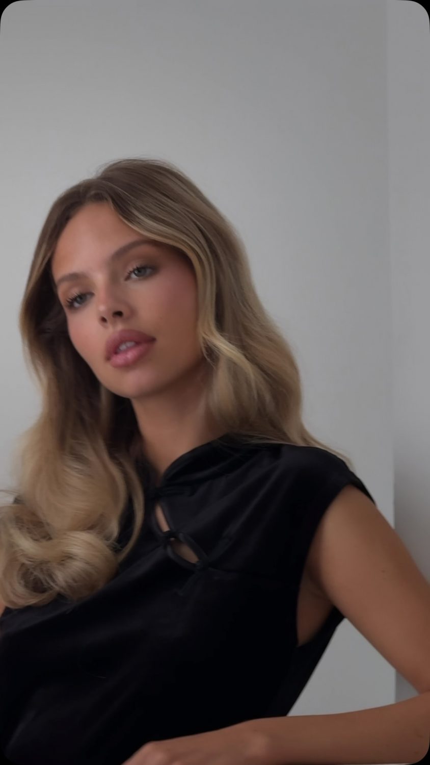 Portrait of a young woman with long wavy blonde hair wearing a black top, looking at the camera.