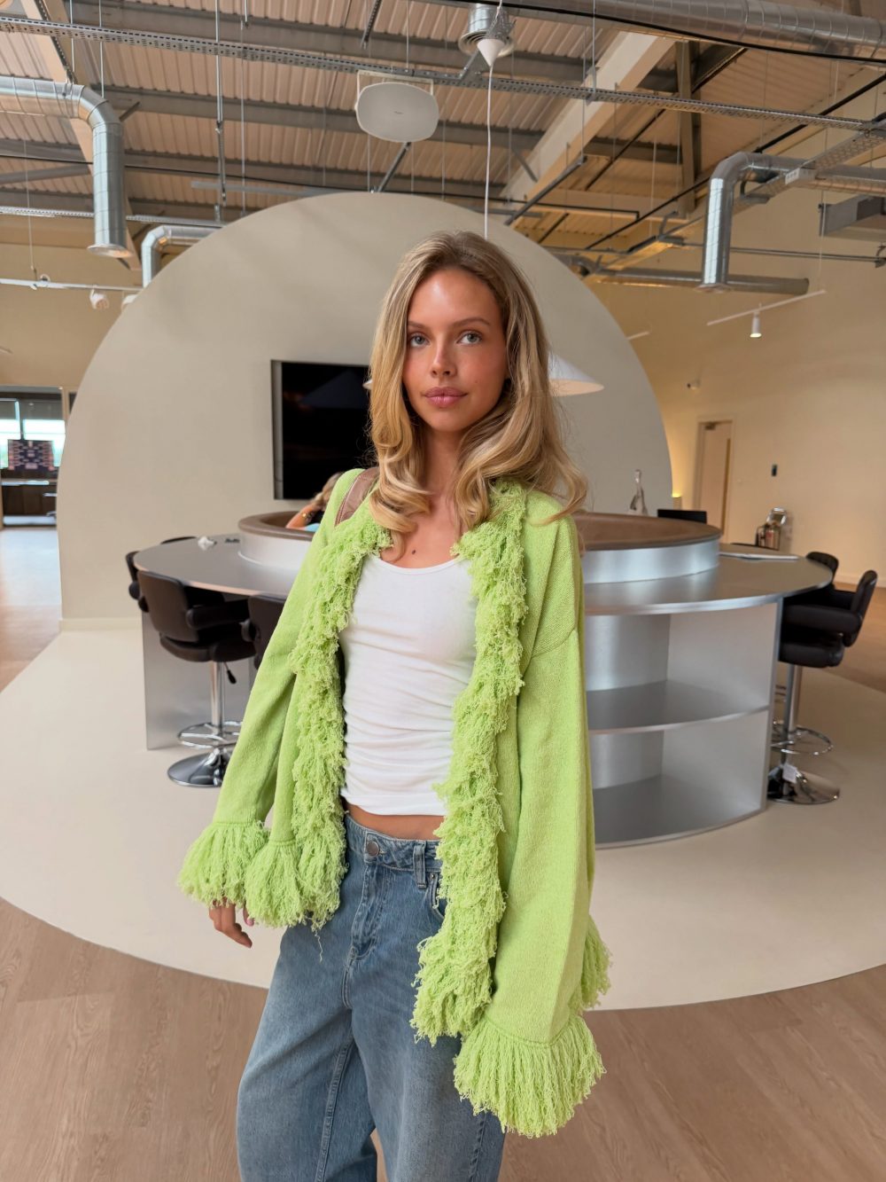 Young woman with blonde hair wearing a lime-green fringed cardigan over a white top and jeans, standing in a modern open-plan lounge with a curved counter in the background.