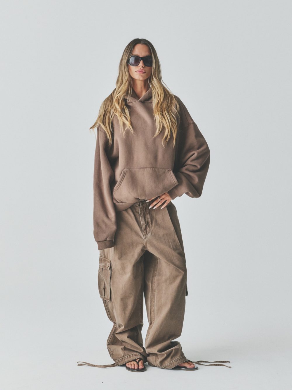 Woman in an oversized brown hoodie and cargo pants wearing black sunglasses, standing against a pale studio backdrop.
