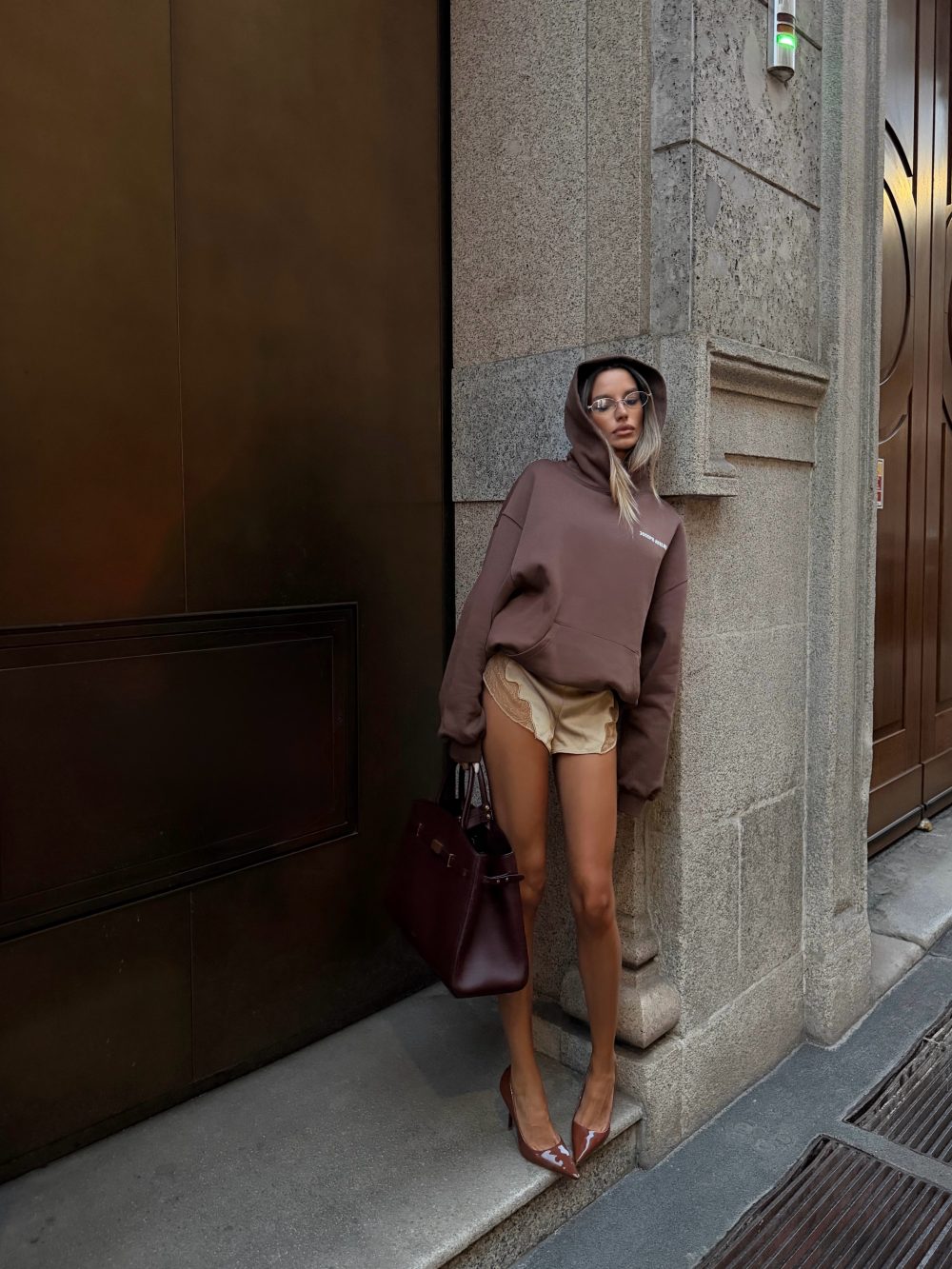 Woman leaning against a stone building in a brown hoodie, beige shorts, and brown heels, carrying a dark bag and wearing round glasses.