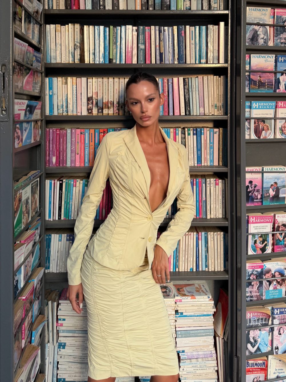 Woman in a pale yellow blazer dress posing in a bookstore aisle with shelves full of colorful magazines around her.