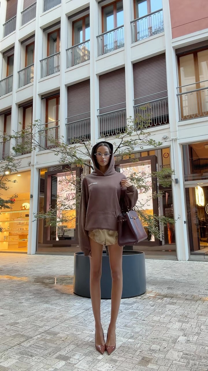 Woman posing in a city plaza wearing a brown hoodie, beige shorts, brown heels, and a purple handbag with a modern storefront backdrop on white-framed buildings.