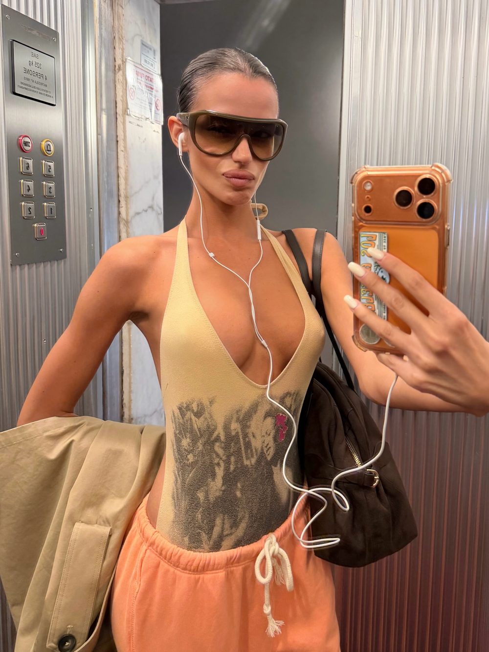 Selfie in an elevator of a person wearing oversized sunglasses, a beige sleeveless top, peach shorts, earphones, and carrying a jacket and bag while holding a gold-colored phone case as they take a photo.