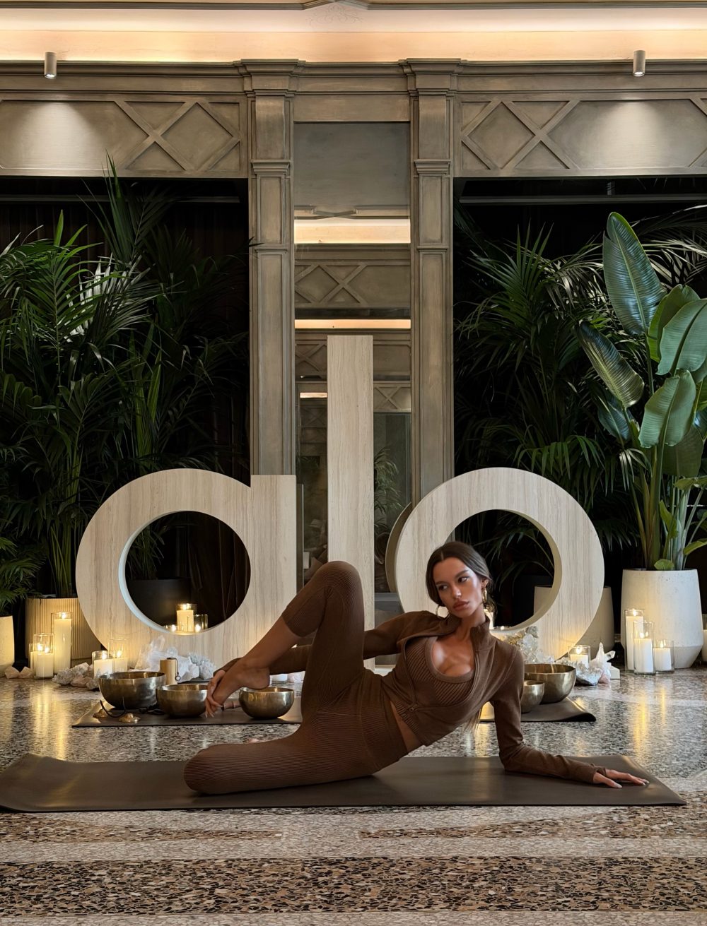 Woman in a brown ribbed outfit performing a yoga pose on a black mat, surrounded by candles and tropical plants in a stylish interior.