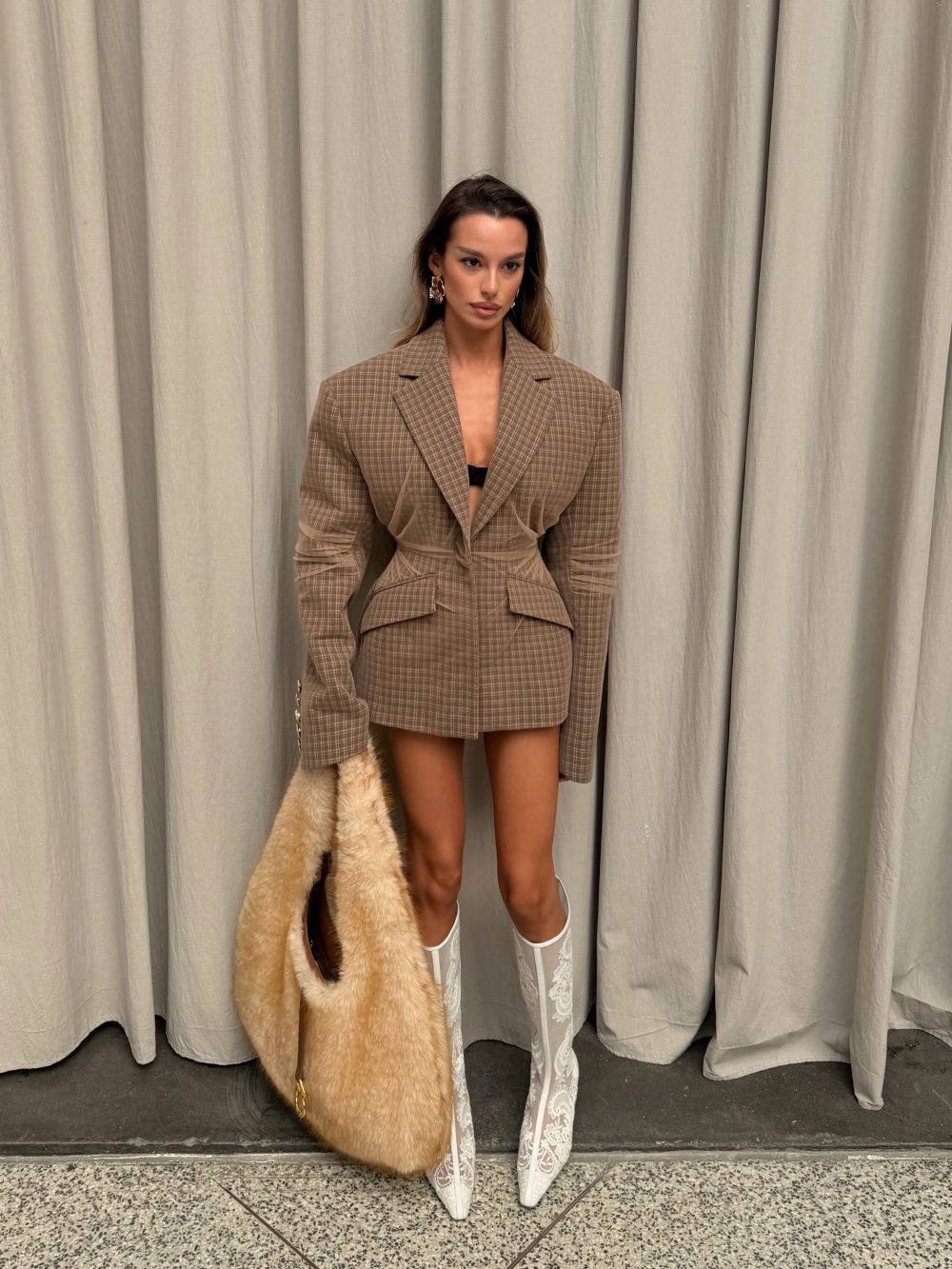 Woman in a brown plaid blazer dress with a black top, holding a beige fur coat, wearing white lace knee-high boots in front of light curtains.