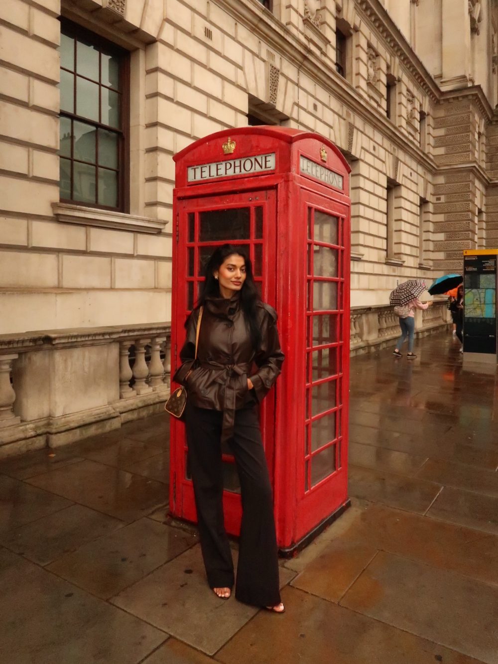 Woman in a brown leather coat posing beside a classic red British telephone booth on a wet city street.