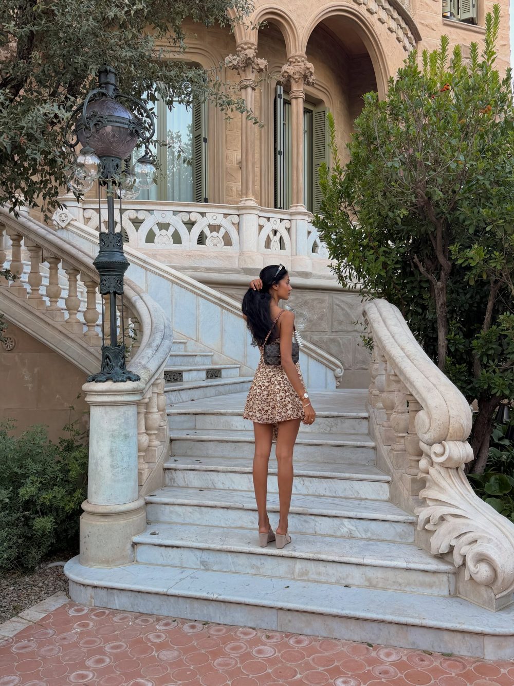 Woman with long dark hair in a leopard print dress stands on an ornate marble staircase outside a beige stone building with arches and greenery.