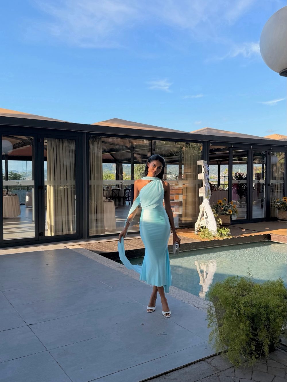 Woman in a mint blue, one-shoulder evening gown standing beside a pool outside a glass-walled venue.