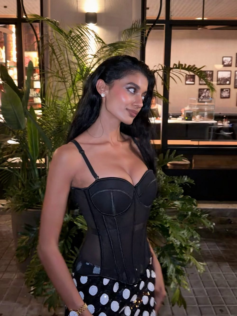 Woman in a black corset top and polka-dot skirt standing among lush plants at night in an urban outdoor setting, facing left.