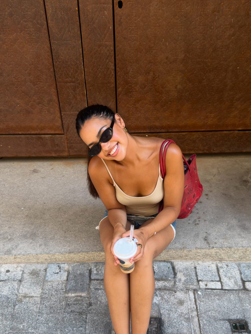 Woman sitting on concrete steps against a rusted wall, wearing sunglasses and a beige tank top, holding a cup with a straw and a red bag nearby.