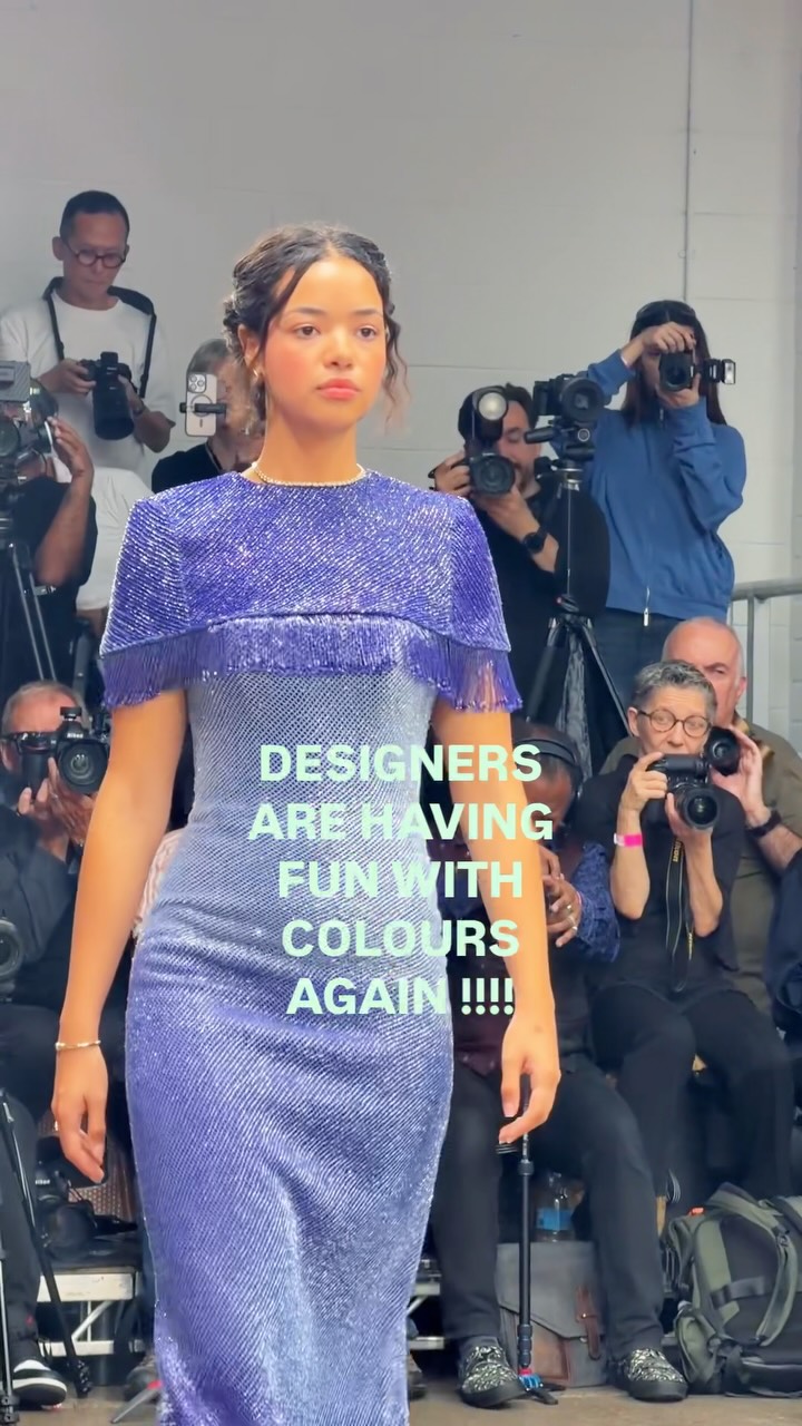Model in a shimmering purple dress with fringe sleeves on a runway; photographers surround her, capturing the moment.