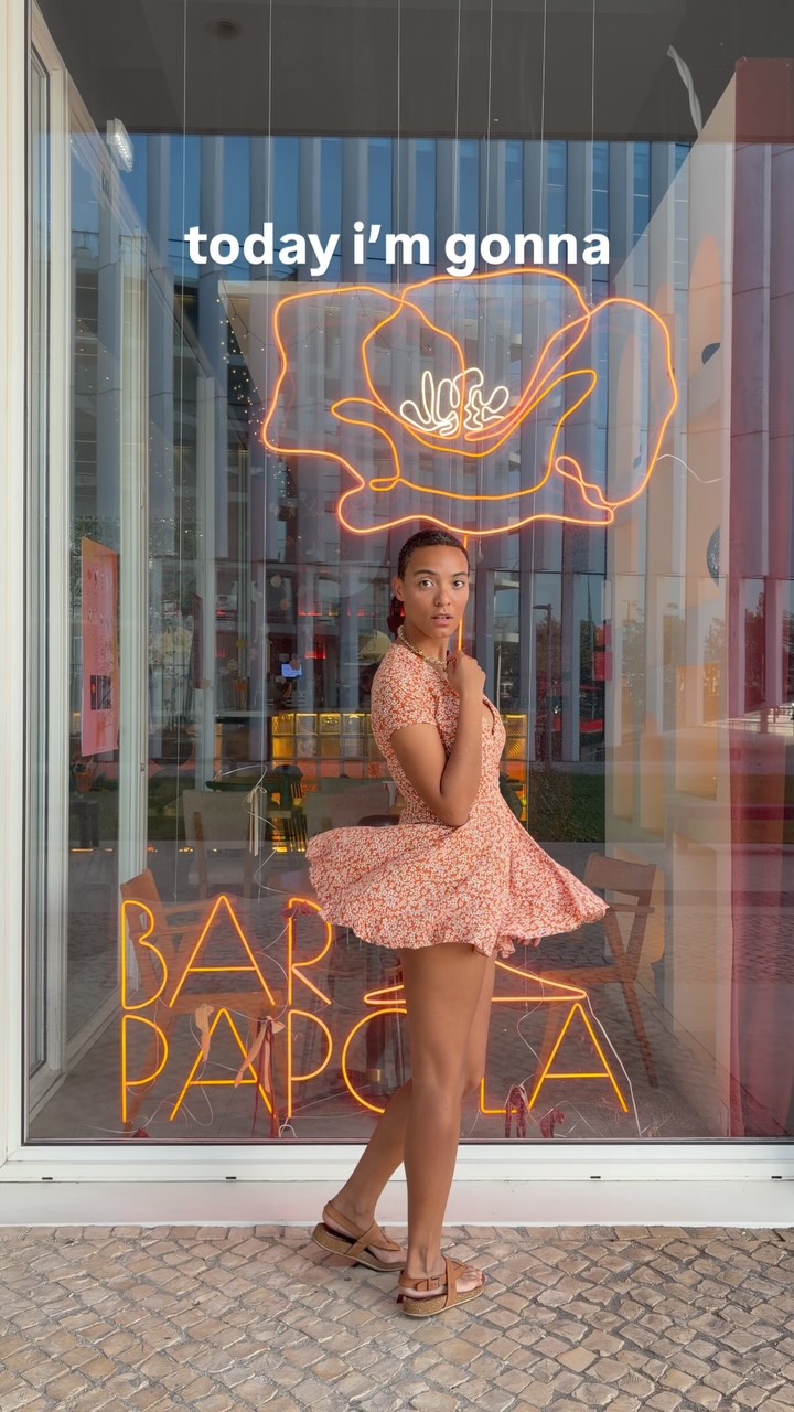 Woman in a peach floral dress poses in a storefront window with orange neon signs reading 'today I'm gonna' and 'BAR' in the background.