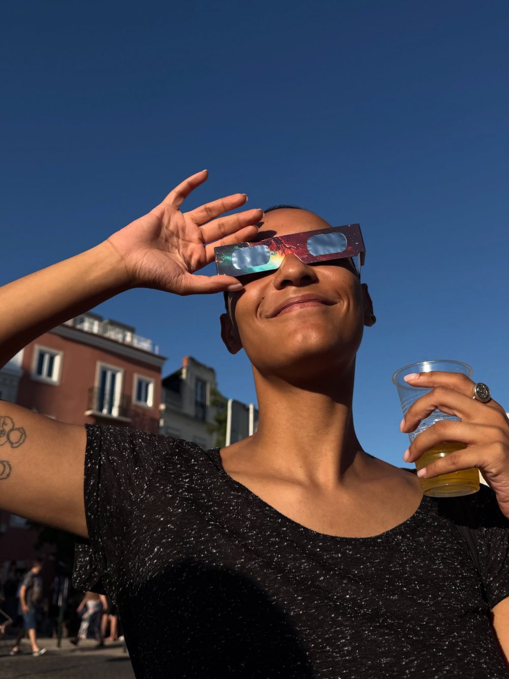 Person wearing galaxy-pattern novelty sunglasses outdoors against a clear blue sky, shielding eyes with a hand and holding a plastic cup with a yellow drink in the other hand, smiling signally.