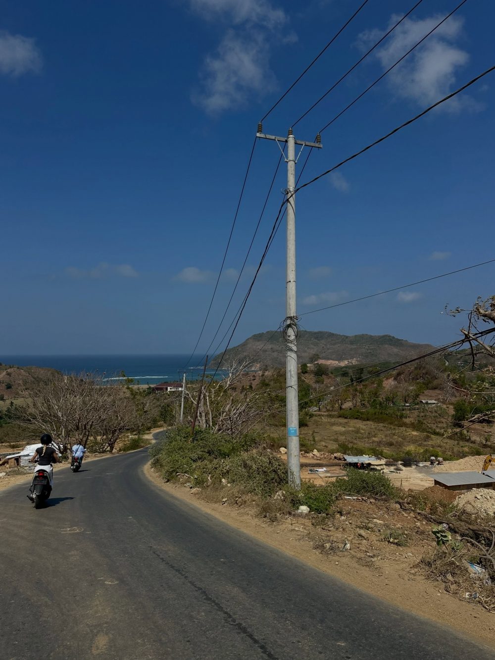 Two people on scooters ride along a winding coastal road with a tall utility pole and wires overhead, mountains and sea in the background.