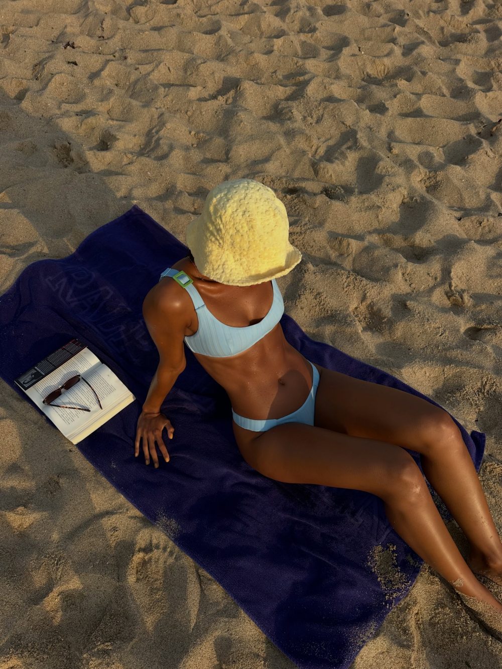 Person sunbathing on a sandy beach, wearing a light blue bikini and a pale yellow wide-brimmed hat, lying on a dark purple towel with an open book and sunglasses nearby.