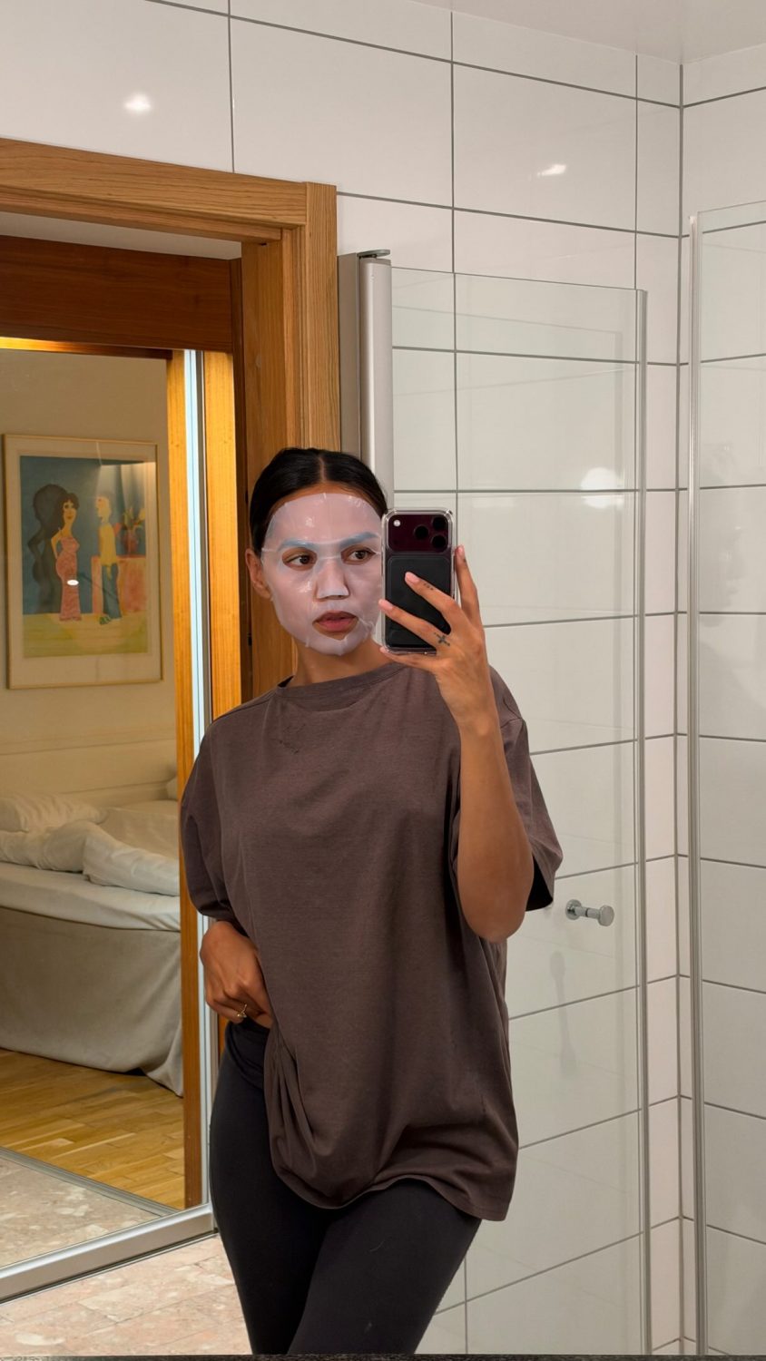 Person wearing a facial sheet mask takes a mirror selfie in a white-tiled bathroom, holding a smartphone.
