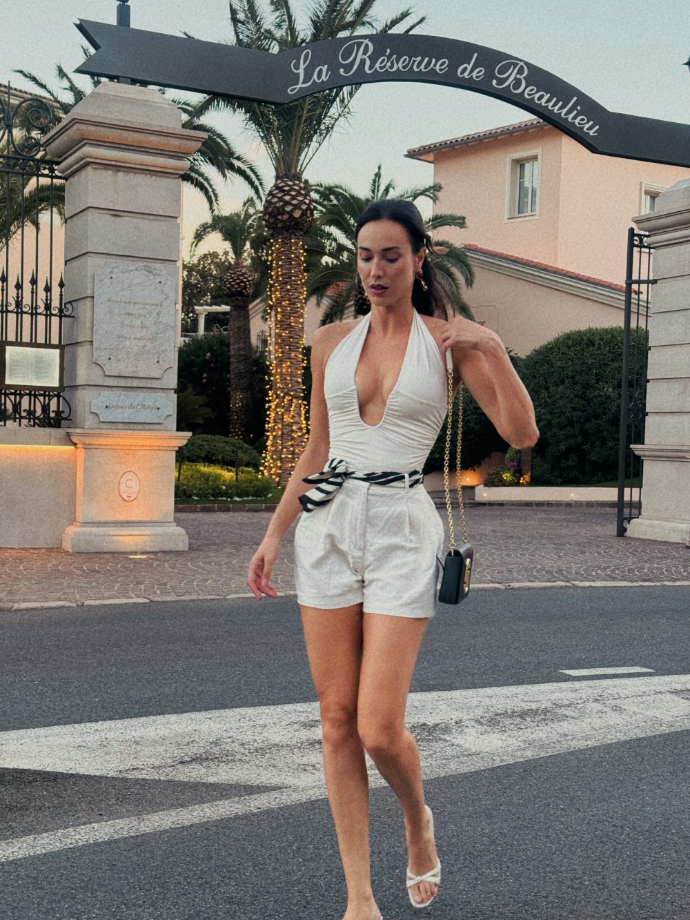 Woman in a white halter romper walking on a street near a gated resort entrance with palm trees and lights in the background.