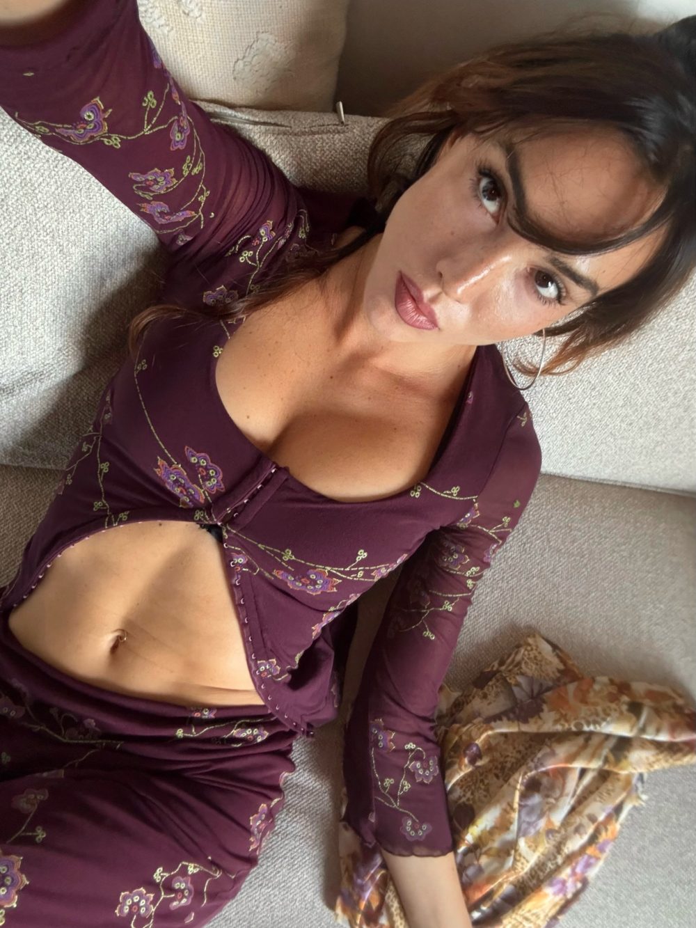 Woman lying on a beige sofa, wearing a maroon floral dress, looking at the camera.
