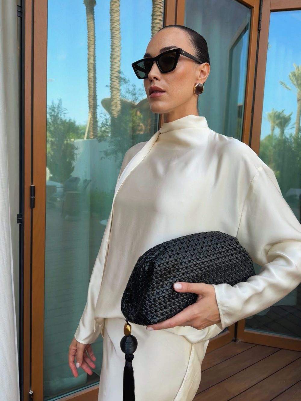 Woman in a cream blouse wearing oversized black sunglasses, holding a textured black clutch outdoors beside glass doors with palm trees in the background.