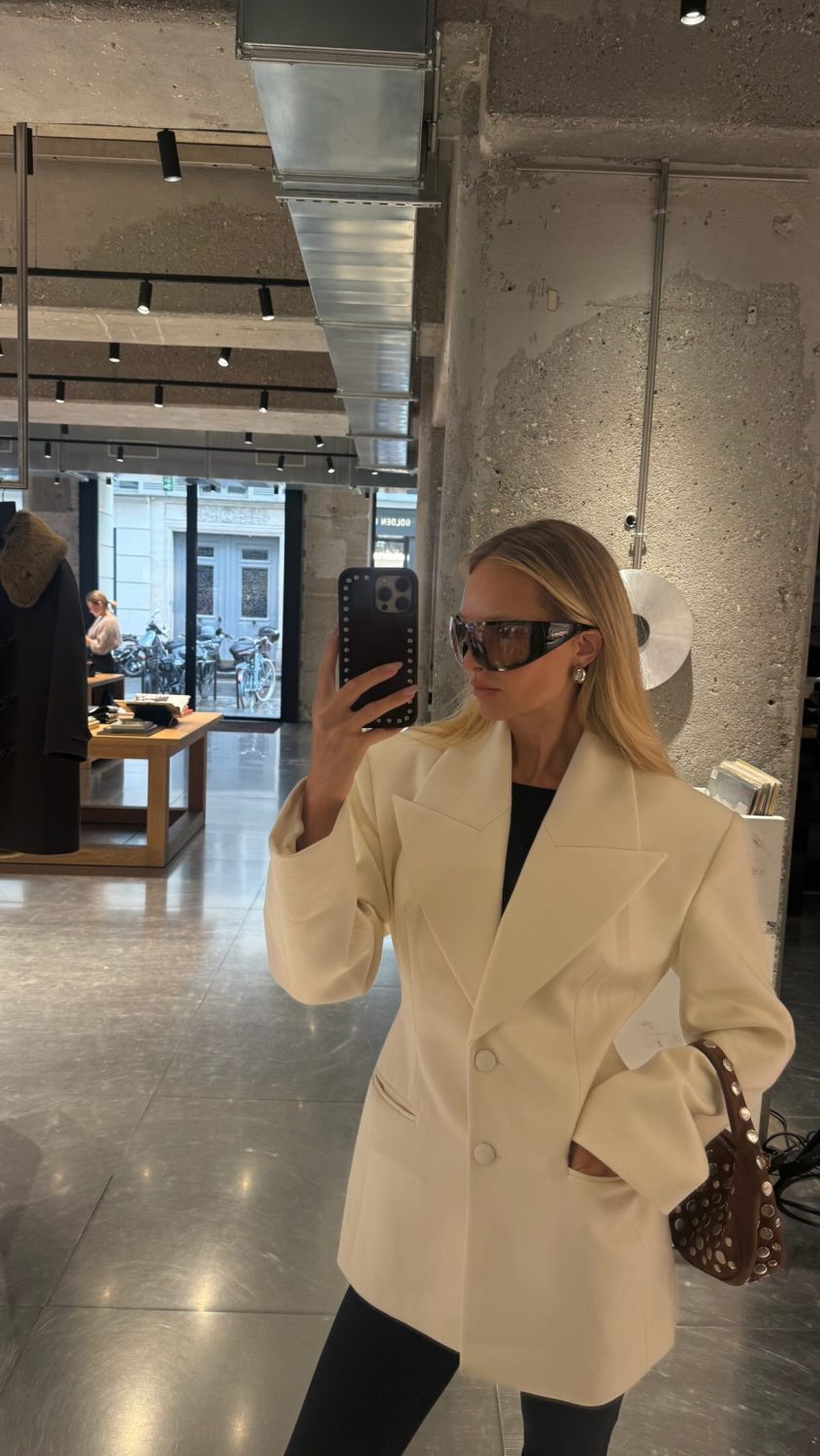 Woman in a cream blazer takes a mirror selfie in a modern retail store with exposed concrete walls and metal ducts, carrying a studded brown handbag.