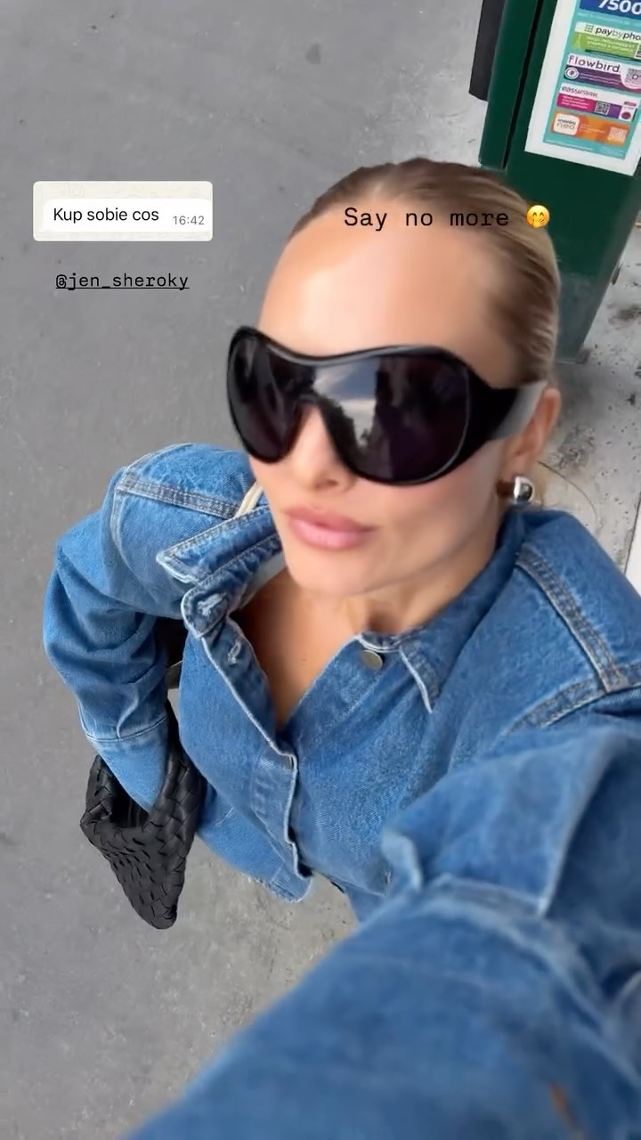 Woman in oversized black sunglasses and a blue denim jacket takes a top-down selfie on a sidewalk, holding a black woven bag.