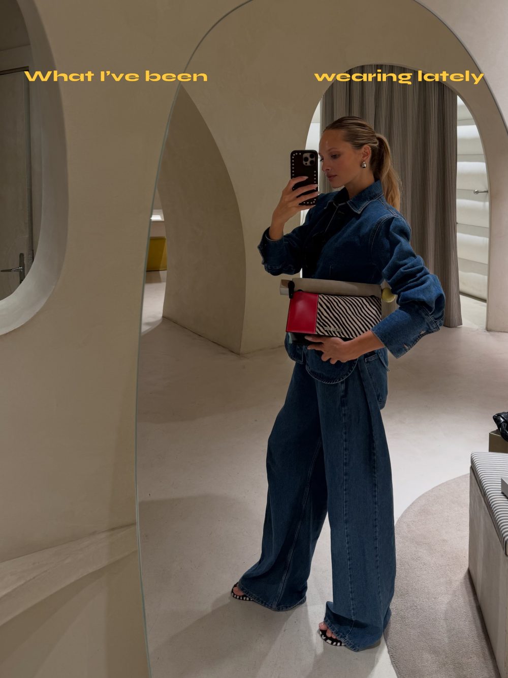 Woman in a denim jumpsuit taking a mirror selfie in a fitting room with curved beige walls and a yellow text overlay: 'What I've been wearing lately'
