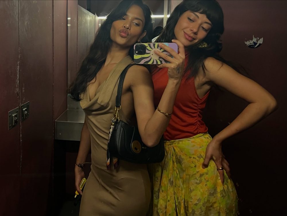 Two women posing for a mirror selfie in an elevator, one in a beige dress and the other in a red top with a floral skirt, holding a patterned phone case.