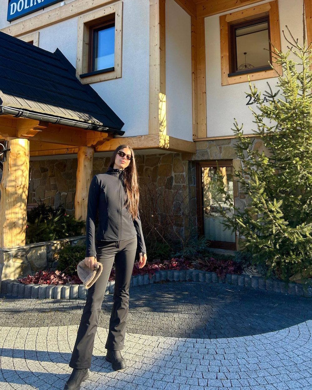 Woman in black athletic outfit standing outside a stone-and-wood building with timber framing and large windows.
