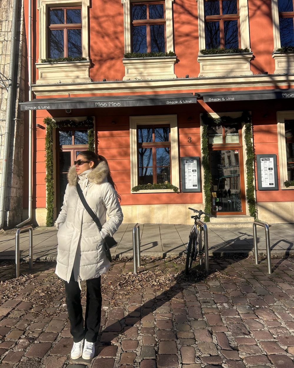 Woman in a white winter coat with a fur-trimmed hood and sunglasses stands on a cobblestone street in front of a coral building with a Moo Moo steak & wine awning.