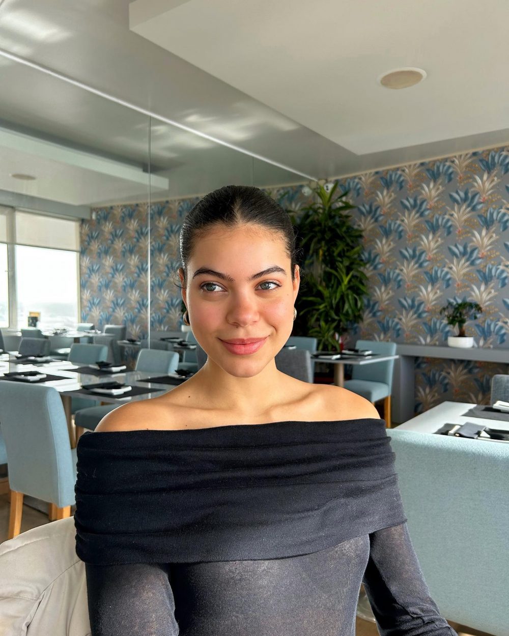 Portrait of a smiling woman with dark hair tied back, wearing a black off‑the‑shoulder top, in a dining room with blue tropical wallpaper.