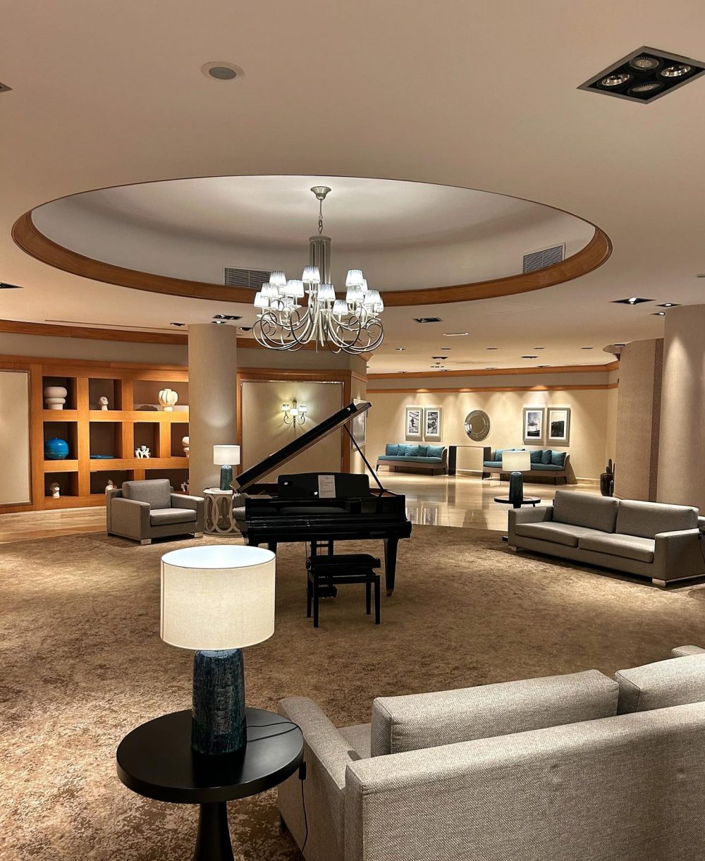Elegant hotel lounge with a grand piano centerpiece, circular chandelier, and beige seating area.