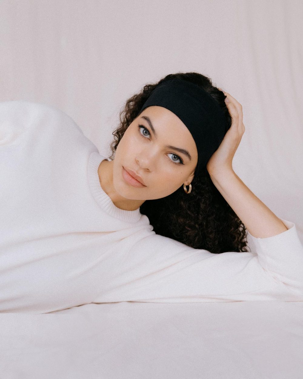 Portrait of a woman with blue-gray eyes wearing a black headband and white sweater, lying on a pale pink surface with her hand in her hair.