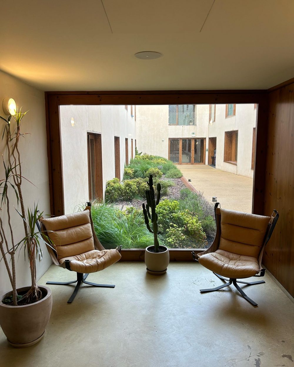 Sunlit seating area with two tan leather lounge chairs facing a large window overlooking a courtyard garden.