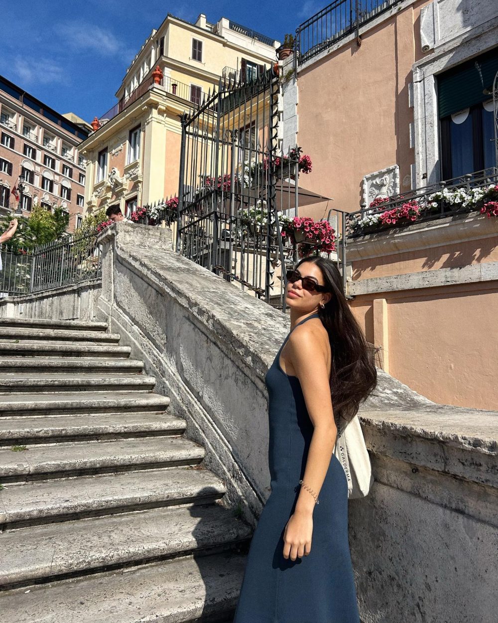 Woman in a blue sleeveless dress and sunglasses walks up a stone staircase beside peach-colored buildings with flower boxes under a clear blue sky.