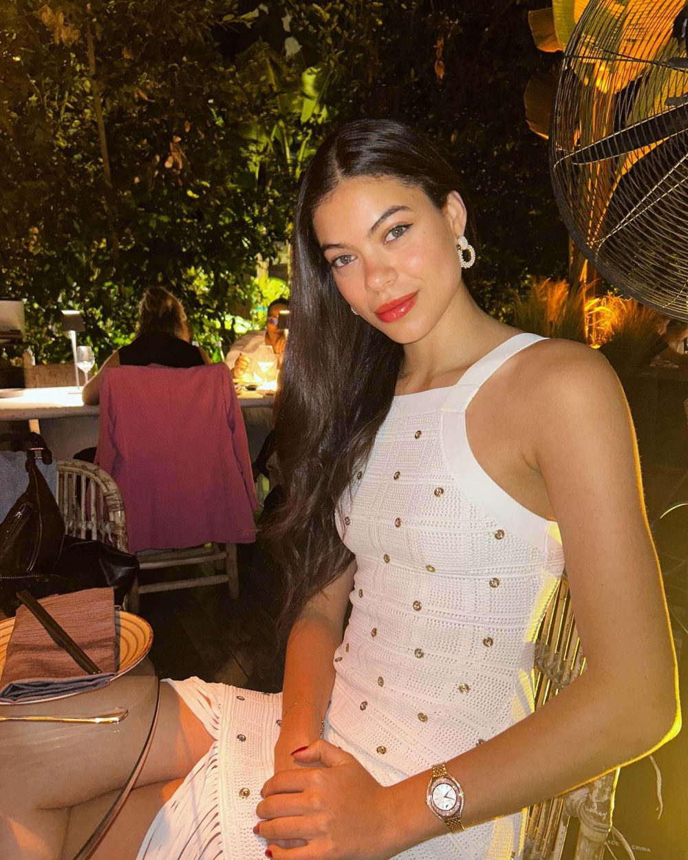 Woman with long dark hair in a white embellished dress sits at an outdoor night restaurant, smiling at the camera.