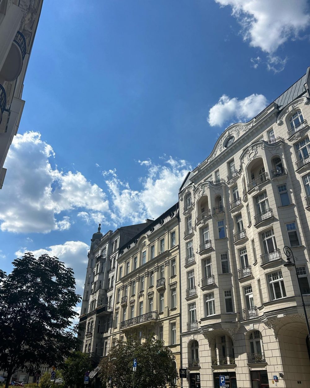 Ornate white apartment building with balconies along a sunny street and a clear blue sky above.