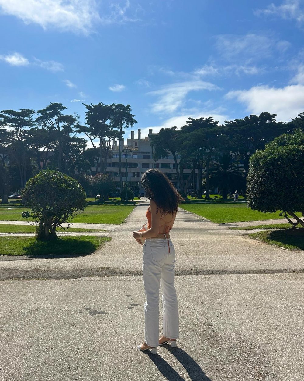 Back view of a woman with curly hair in a peach top and white jeans standing on a park path, with trees and a building in the background.