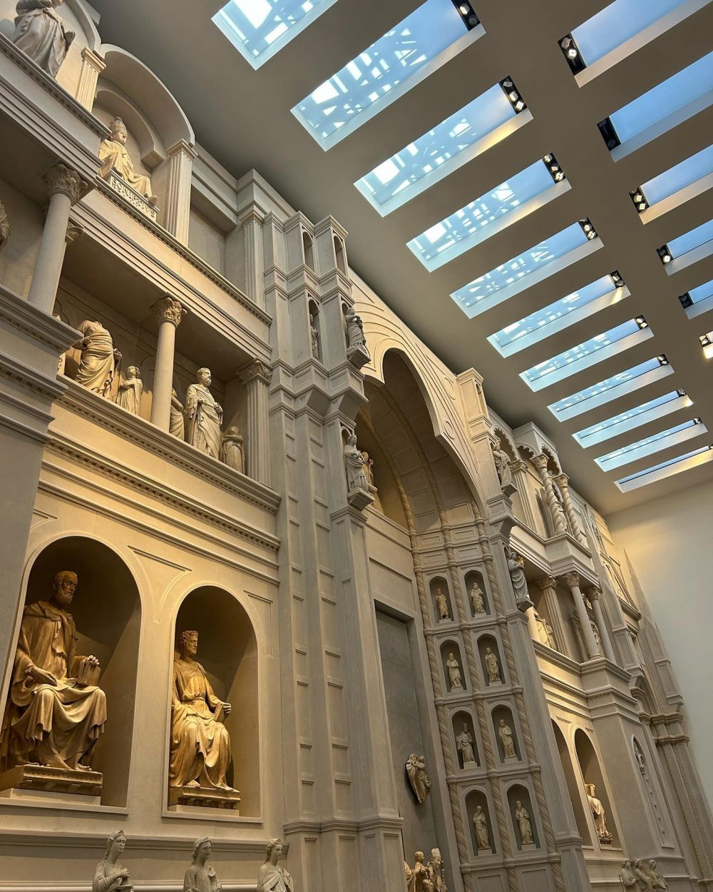 White marble classical sculpture gallery with statues displayed in niches along a tall, ornate wall of arches and columns.