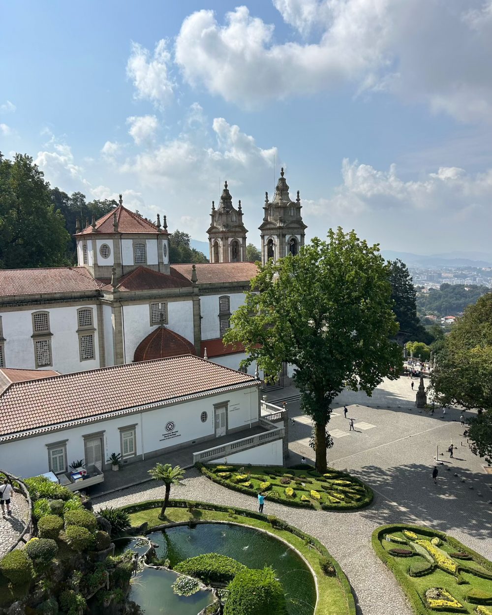 Historic white building with red-tiled roofs, ornate twin bell towers, and a formal garden with circular ponds in the foreground.