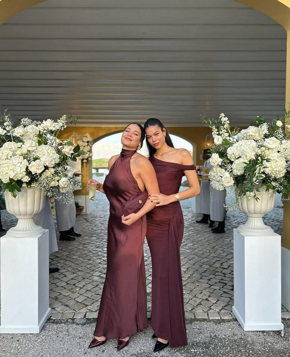 Two women in burgundy gowns pose together under a decorated arch, holding a clutch and a drink amid white floral arrangements at a formal event.