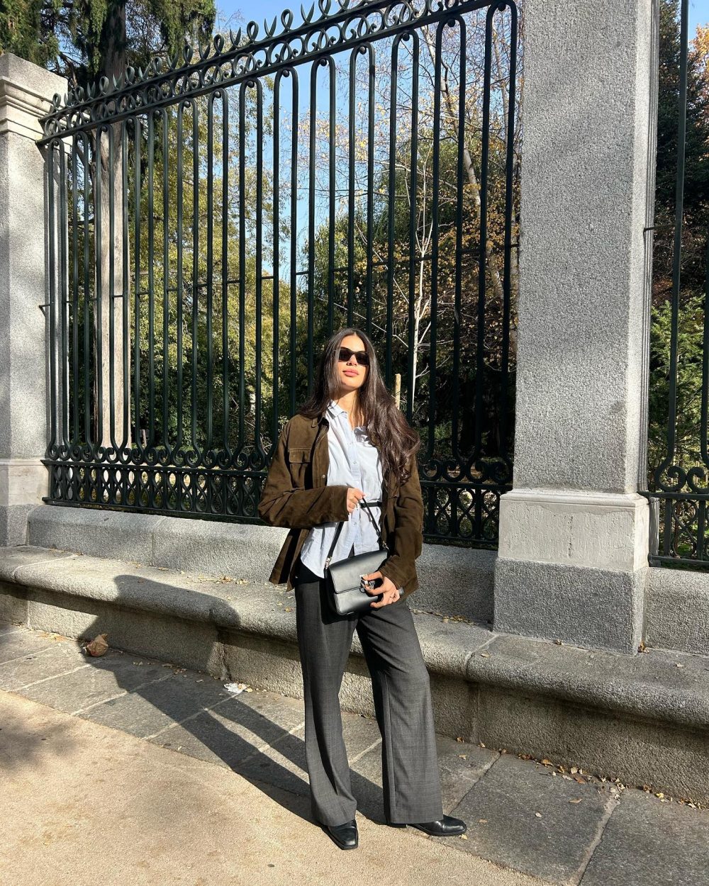 Woman standing in front of an ornate iron fence, wearing sunglasses, a white shirt, brown jacket, dark trousers, and carrying a black handbag.