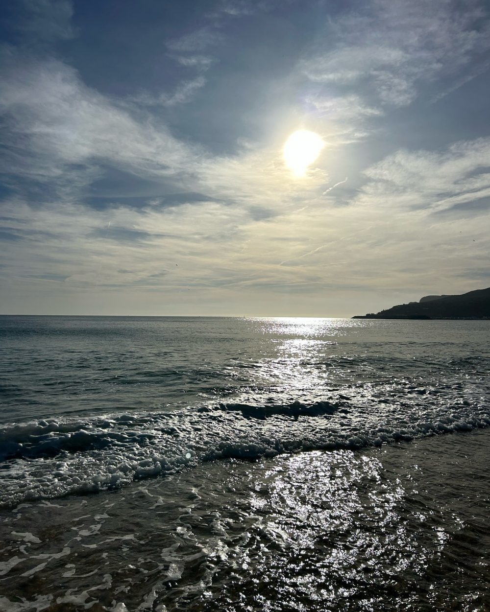 Sunlit sea with shimmering water and a distant headland on the right, under a blue sky with wispy clouds.
