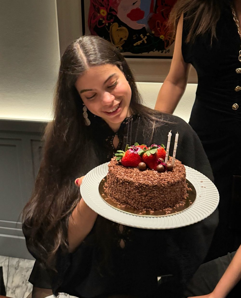 Woman in a black top holds a chocolate cake topped with strawberries and two candles, smiling at the celebration party edgewise.