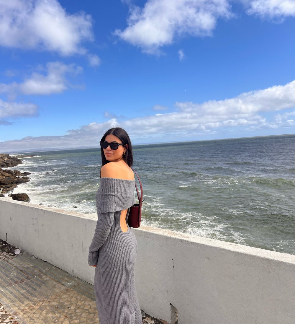 Woman in a gray off-the-shoulder knit dress and sunglasses stands along a seaside concrete barrier, looking toward the camera.