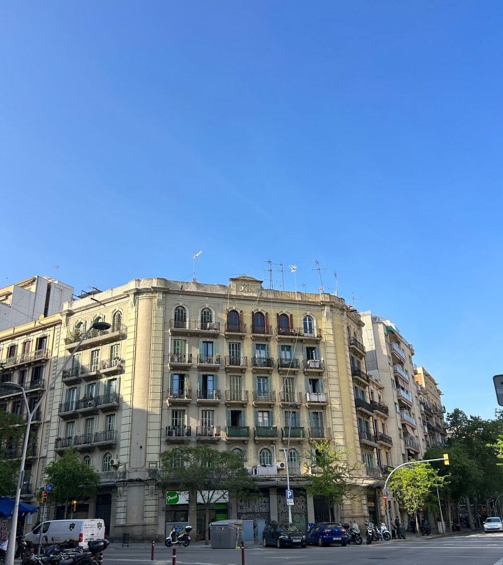 Beige European-style apartment building with wrought-iron balconies on a sunny street, against a clear blue sky.