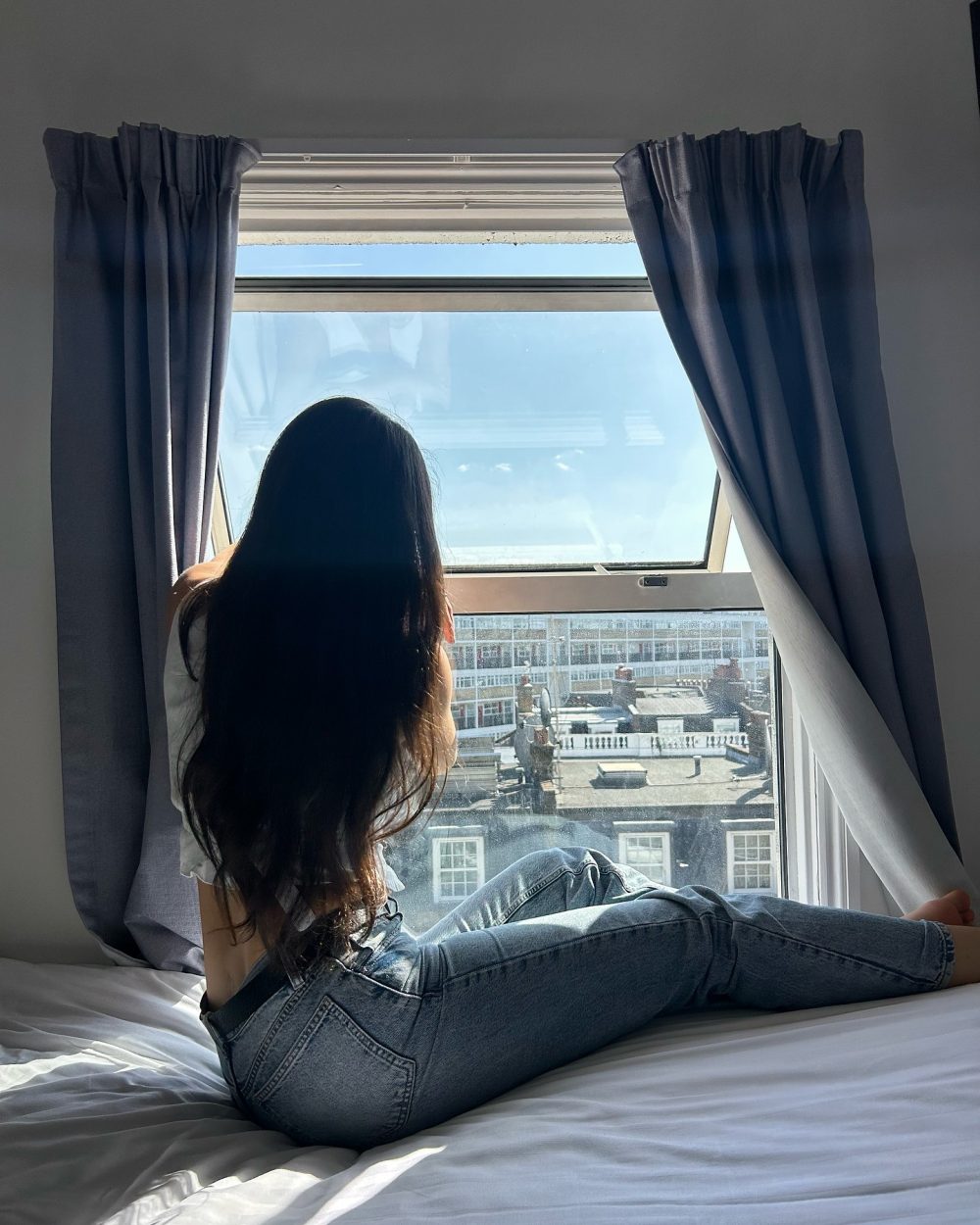 Person with long dark hair in jeans sits on a bed, looking out a large window with blue curtains.