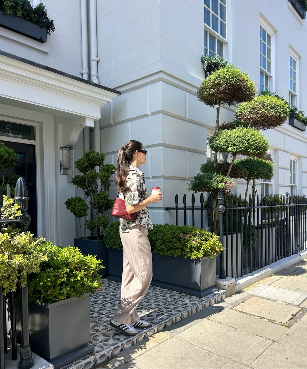 Woman with a coffee cup standing on patterned steps outside a white townhouse with sculpted topiary trees and a black iron fence