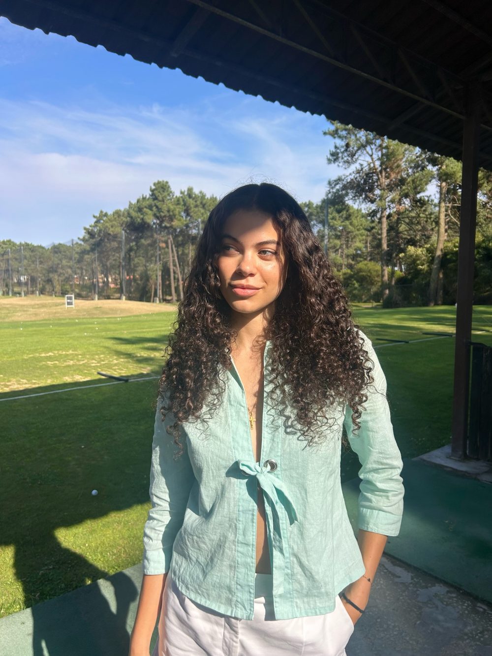 Young woman with curly hair standing outside in a light blue buttoned top and white pants, under a shaded structure, on a sunny day.