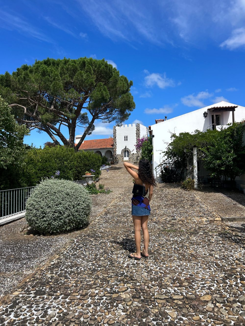 Woman standing on a pebble path in a sunny courtyard with white buildings and a large pine tree behind her.