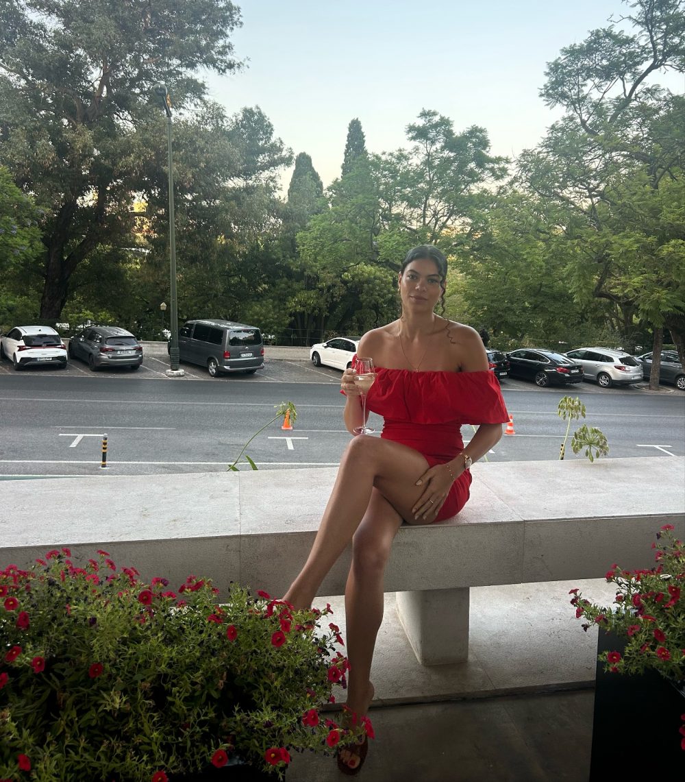 Woman in a red off-shoulder dress sits on a concrete ledge, holding a wine glass, with a street and trees in the background.